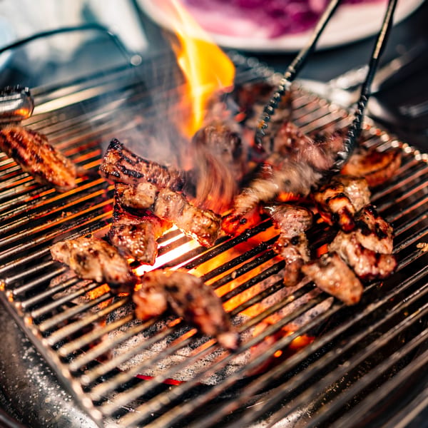 Grilled meat cooking over open flames on a barbecue with tongs turning the pieces.